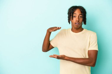 Genç Afrikalı Amerikalı bir adam mavi arka planda izole edilmiş. Elleri arasında bir kopya alanı tutarken şok olmuş ve hayrete düşmüştü..