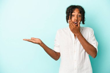 Mavi arka planda izole edilmiş genç Afro-Amerikalı bir adam avuç içinde fotokopi alanı tutuyor, el üstünde tutuyor. Şaşırmış ve mutlu..