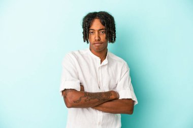Mavi arka planda izole edilmiş, hoşnutsuzca kaşlarını çatmış, kollarını kavuşturmuş genç Afrikalı Amerikalı bir adam..