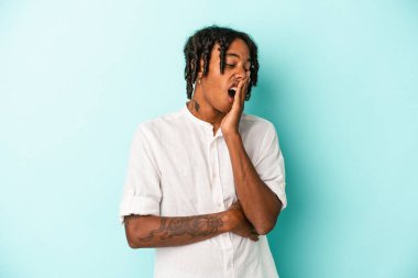 Genç Afrikalı Amerikalı bir adam mavi arka planda izole edilmiş esniyor ve ağzı eliyle kapalı yorgun bir jest yapıyor..