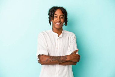 Genç Afro-Amerikalı, mavi arka planda izole edilmiş, kendinden emin, kararlılıkla kollarını kavuşturuyor..