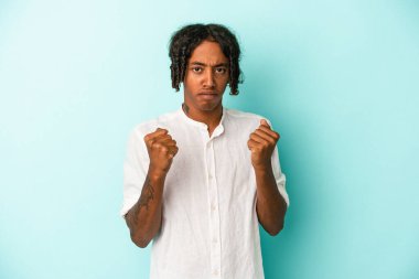 Mavi arka planda izole edilmiş genç Afro-Amerikalı adam yumruğunu kameraya gösteriyor, agresif yüz ifadesi..