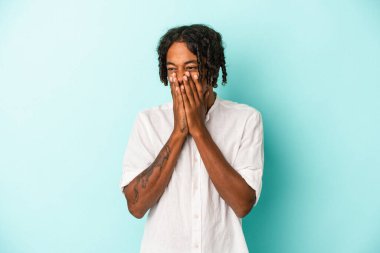 Genç Afrikalı Amerikalı bir adam mavi arka planda izole edilmiş bir şekilde bir şeylere gülüyor, elleriyle ağzını kapatıyor..