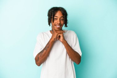 Mavi arka planda izole edilmiş genç Afrikalı Amerikalı ellerini çenesinin altında tutuyor ve mutlu bir şekilde yan tarafa bakıyor..