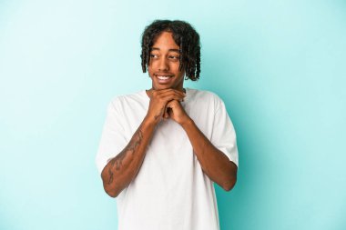 Genç Afro-Amerikalı adam mavi arka planda izole edilmiş şans için dua ediyor, şaşırmış ve ağzını açıyor..