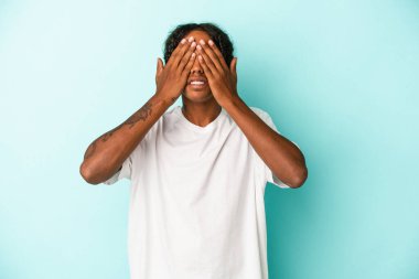Genç Afrikalı Amerikalı bir adam mavi arka planda izole edilmiş. Gözleri elleriyle kapatmaktan korkuyor..