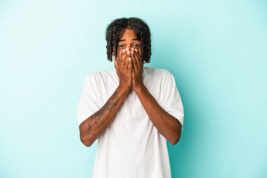 Genç Afrikalı Amerikalı bir adam mavi arka planda izole edilmiş. Şok olmuş, ağzı ellerle kaplanmış, yeni bir şey keşfetmeye hevesli..