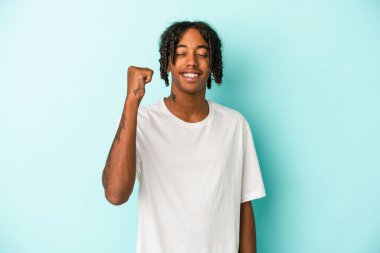 Genç Afrikalı Amerikalı bir adam mavi arka planda izole edilmiş zafer, tutku ve coşkuyu kutluyor, mutlu bir ifade..