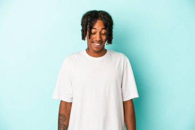 Genç Afrikalı Amerikalı bir adam mavi arka planda izole edilmiş, güler ve gözlerini kapatır, rahatlamış ve mutlu hisseder..