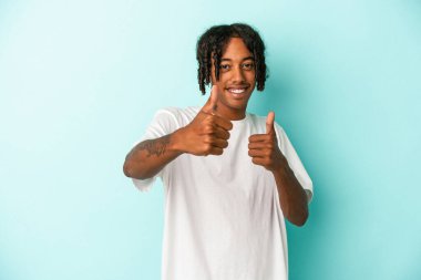Genç Afrikalı Amerikalı adam mavi arka planda izole edilmiş. Baş parmağını kaldırıyor, gülümsüyor ve kendinden emin..