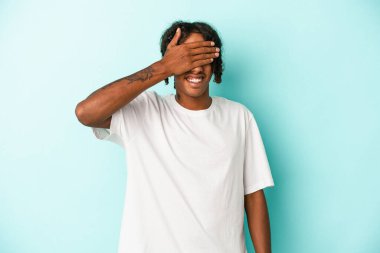 Mavi arka planda izole edilmiş genç bir Afro-Amerikan erkeği elleriyle gözlerini kapatıyor, gülümseyerek büyük bir sürpriz bekliyor..