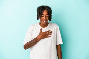 Mavi arka planda izole edilmiş genç Afro-Amerikalı adam yüksek sesle gülüyor ve elini göğsüne koyuyor..