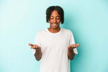 Genç Afrikalı Amerikalı adam mavi arka planda izole edilmiş. Elinde avuç içi olan bir şey var. Kameraya teklif ediyor..