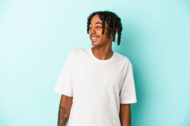 Genç Afrikalı Amerikalı adam mavi arka planda izole edilmiş rahat ve mutlu bir şekilde gülüyor, boynunda dişlerini gösteriyor..