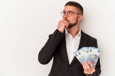Beyaz arka planda soyutlanmış, şüpheli ve şüpheci bir ifadeyle yan bakan beyaz banknotlar tutan genç bir beyaz iş adamı..