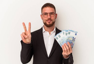 Beyaz arka planda iki numarayı parmaklarıyla gösteren banknotları tutan genç beyaz bir adam..