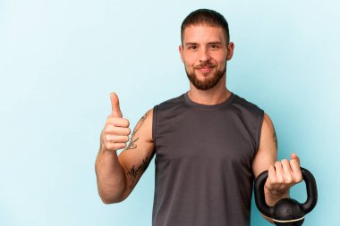 Ketlebell 'i tutan genç beyaz adam mavi arka planda izole edilmiş gülümsüyor ve başparmağını kaldırıyor.