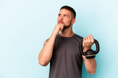 Ketlebell 'i tutan genç beyaz adam mavi arka planda izole edilmiş şüpheli ve şüpheci bir ifadeyle yana bakıyor..