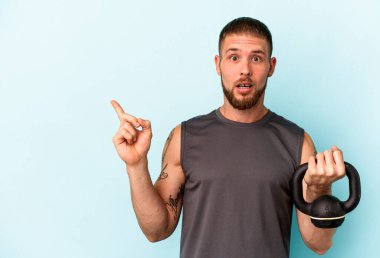 Ketlebell 'i tutan genç beyaz adam mavi arka planda izole edilmiş, yan tarafı işaret ediyor.