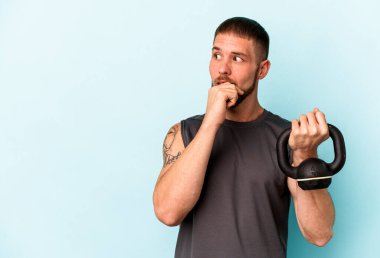 Ketlebell 'i tutan genç beyaz adam mavi arka planda izole edilmiş. Bir şeyin fotokopi odasına baktığını düşünüyor..
