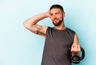 Ketlebell 'i tutan genç beyaz adam mavi arka planda izole edilmiş kafasının arkasına dokunuyor, düşünüyor ve bir seçim yapıyor..