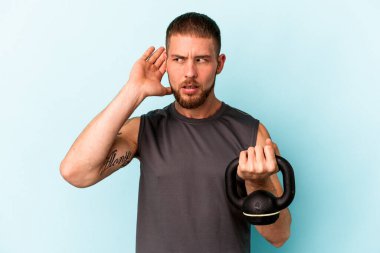 Ketlebell 'i tutan beyaz bir genç mavi arka planda izole edilmiş bir şekilde bir dedikoduyu dinlemeye çalışıyor..
