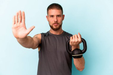 Ketlebell 'i tutan genç beyaz adam mavi arka planda izole edilmiş, elini uzatmış, dur işareti göstererek seni engelliyor..