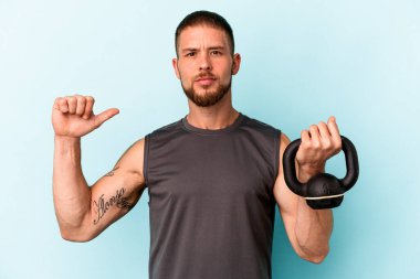 Ketlebell 'i mavi arka planda tutan genç beyaz adam gururlu ve kendinden emin hissediyor, örnek olarak..