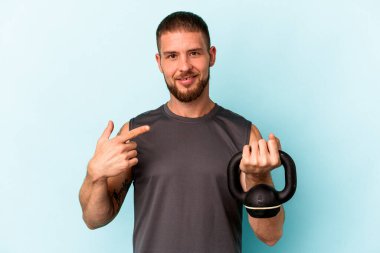 Ketlebell 'i tutan genç beyaz adam mavi arka planda tek başına gömlek fotokopi alanını gösteren gururlu ve kendinden emin birini işaret ediyor.