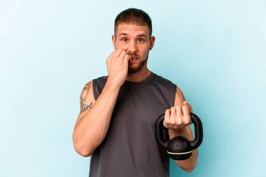 Ketlebell 'i tutan genç beyaz adam mavi arka planda izole edilmiş tırnak yiyor, gergin ve çok endişeli..