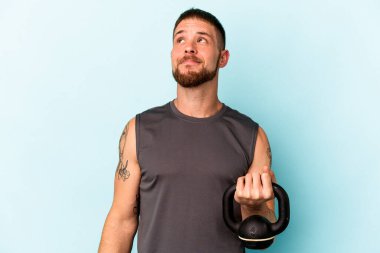 Ketlebell 'i elinde tutan Kafkasyalı genç adam mavi arka planda izole edilmiş hedeflerine ve amaçlarına ulaşma hayali kuruyor.