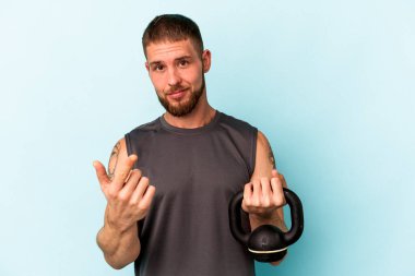 Ketlebell 'i tutan genç beyaz bir adam mavi arka planda izole edilmiş ve sanki seni davet ediyormuş gibi parmağıyla işaret ediyor..