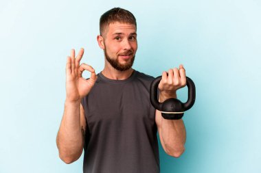 Ketlebell 'i tutan genç beyaz adam mavi arka planda izole edildi. Neşeli ve kendinden emin..
