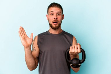 Ketlebell 'i tutan genç beyaz adam mavi arka planda izole edilmiş. Şaşırmış ve şok olmuş..