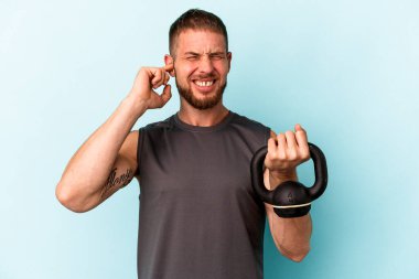Ketlebell 'i tutan genç beyaz adam mavi arka planda izole edilmiş kulakları elleriyle kapatıyor..