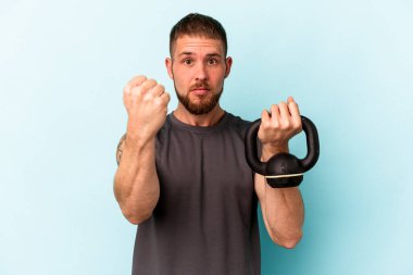 Ketlebell 'i tutan genç beyaz adam mavi arka planda izole edilmiş kameraya yumruğunu gösteriyor, agresif yüz ifadesi var..