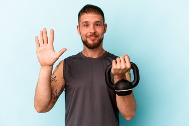 Ketlebell 'i tutan genç beyaz adam mavi arka planda izole edilmiş. Beş numarayı parmaklarıyla gülümsüyor..