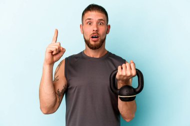 Ketlebell 'i tutan genç beyaz adam mavi arka planda izole edilmiş. Harika bir fikri var, yaratıcılık kavramı..