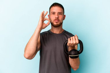 Ketlebell 'i tutan beyaz bir genç mavi arka planda izole edilmiş dudaklarında parmakları olan bir sır saklıyor..