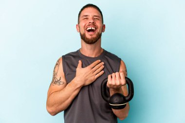 Ketlebell 'i tutan beyaz bir genç mavi arka planda izole edilmiş olarak kahkahalar atıyor..
