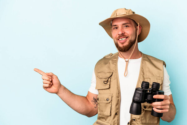 Young caucasian man looking at animals through binoculars isolated on blue background smiling and pointing aside, showing something at blank space.