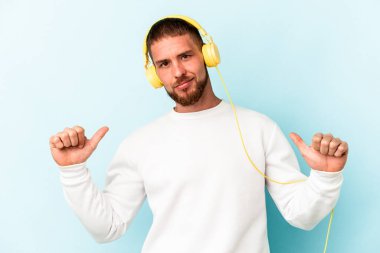Mavi arka planda izole edilmiş müzik dinleyen genç beyaz adam gururlu ve kendinden emin hissediyor, örnek olarak..