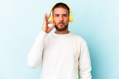 Genç beyaz adam mavi arka planda izole edilmiş müzik dinliyor. Parmakları dudaklarında bir sır saklıyor..
