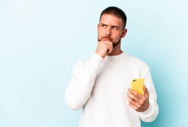 Elinde cep telefonu olan genç beyaz adam mavi arka planda izole edilmiş şüpheli ve şüpheci bir ifadeyle yana bakıyor..