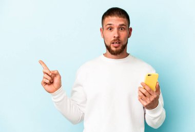 Elinde cep telefonu olan beyaz bir adam mavi arka planda izole edilmiş ve yan tarafı işaret ediyor.