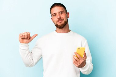 Mavi arka planda cep telefonu tutan genç beyaz adam gururlu ve kendinden emin hissediyor, örnek olarak..
