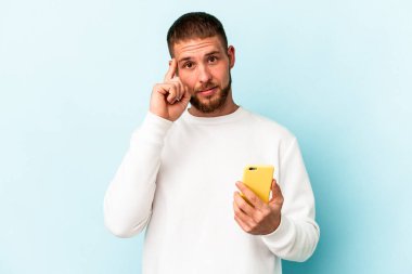 Elinde cep telefonu olan beyaz bir adam mavi arka planda izole edilmiş. Şakağı işaret ediyor, düşünüyor ve bir göreve odaklanıyor..