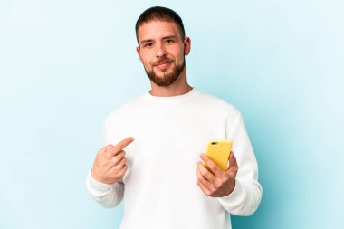Elinde cep telefonu olan beyaz bir adam mavi arka planda tek başına gömlek fotokopi alanını işaret ediyor, gururlu ve kendinden emin.