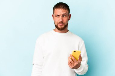 Mavi arka planda cep telefonu tutan genç beyaz adam kafası karışmış ve kendinden emin değil..