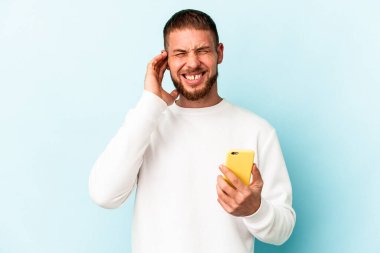 Elinde cep telefonu olan beyaz bir adam mavi arka planda izole edilmiş kulakları elleriyle kapatıyor..